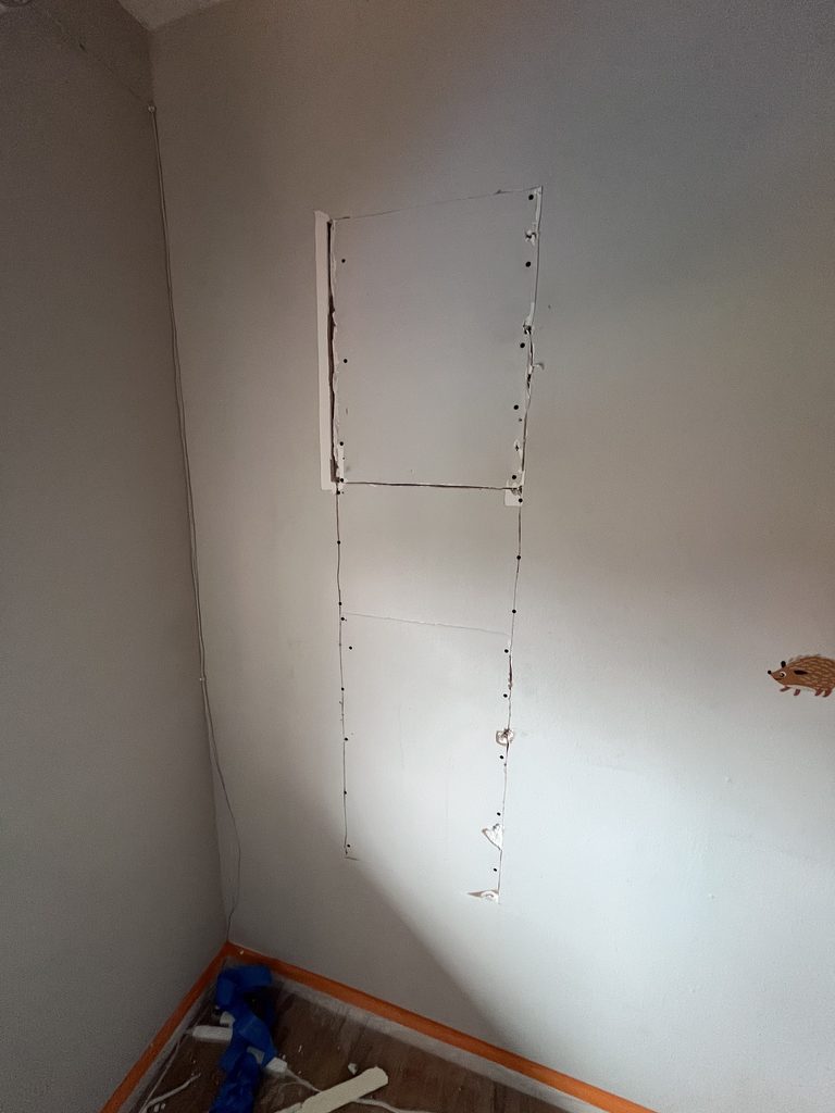 Bedroom wall with a tall rectangular drywall access cut, screws and untaped seams visible, before finishing