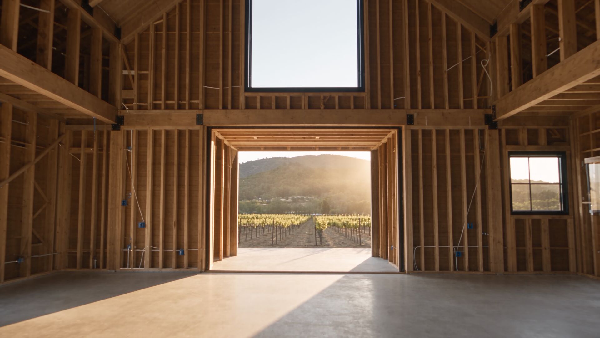 Inside a Wine Country barn at the framing stage: exposed wood studs and rafters, a concrete floor, and a vineyard and hills visible through the open doorway